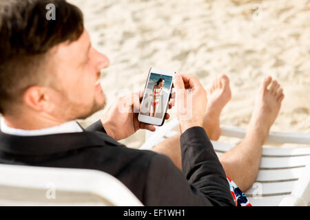 Businessman holding portable avec une photo de votre petite amie assis sur le transat habillé en costume et courts-circuits sur la plage Banque D'Images