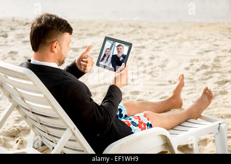 Businessman habillés en costume et pantalon ayant appel vidéo avec tablette numérique sur le transat à la plage Banque D'Images