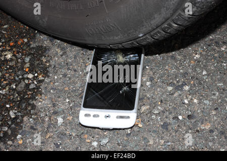 Un livre blanc HTC téléphone avec un écran tactile brisé sur une chaussée en vertu d'un pneu de voiture. Banque D'Images