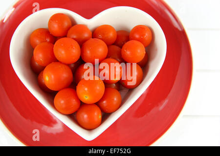 Close up de tomates cerise rouge vif dans un bol en forme de coeur sur plaque rouge sur un fond de bois blanc. Banque D'Images