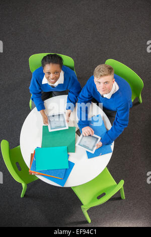 Vue de dessus de teenage boy and girl sitting at table ronde avec les tablettes numériques, looking at camera