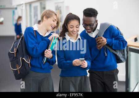 Trois smiling students wearing blue l'uniforme scolaire avec un téléphone mobile dans le corridor de l'école Banque D'Images