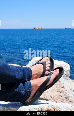 Gros plan des pieds d'un homme avec des tongs qui est relaxant près de l'océan dans l'été Banque D'Images