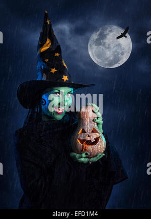 Sorcière avec peau verte tenue Halloween citrouille sculptée à lune et ciel noir avec de la pluie Banque D'Images