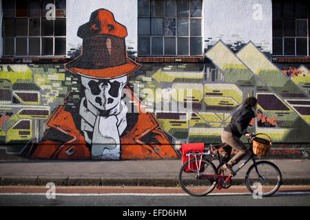 Les cycles d'une femme passé un morceau de Urban street art et graffiti sur le côté d'un immeuble à Stokes Croft, Bristol. Banque D'Images