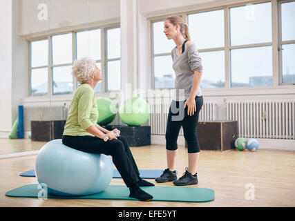 Thérapeute physique de mandater un senior woman at rehab. Parler avec l'ancien entraîneur féminin woman sitting on fitness ball holding du Banque D'Images