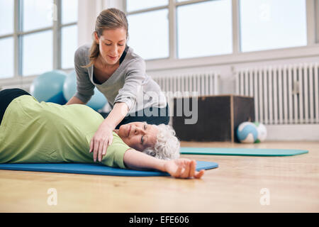 Les femmes âgées se trouvant sur tapis d'exercice doing stretching entraînement pour les muscles du dos avec l'entraîneur de l'aide. Femme trainer helping elder Banque D'Images