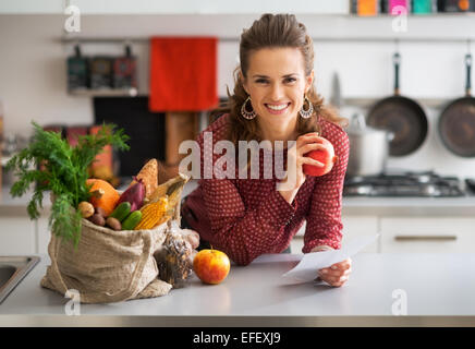 Portrait of happy young housewife holding grocery shopping vérifie dans la cuisine Banque D'Images