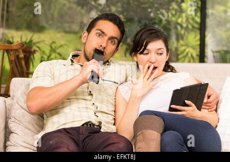 Cute couple cuddling surpris dans le canapé tout en regardant la télévision et à l'aide de tablet Banque D'Images