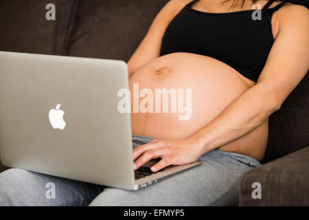 Baby bump, image de 8 mois femme enceinte assise sur un canapé, avec un imac sur ses genoux, la dactylographie. Banque D'Images