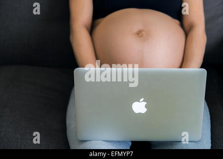 Baby bump, image de 8 mois femme enceinte assise sur un canapé, avec un iMac sur ses genoux, la dactylographie. Banque D'Images