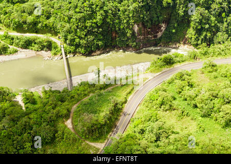 Une perspective aérienne montre une route courbe à deux voies longeant une rivière verdoyante et boueuse à travers une forêt dense et verdoyante. La rivière coule à travers une vallée étroite avec des rives rocheuses, et une grande Banque D'Images
