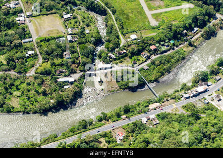 Une photographie aérienne montre une rivière boueuse serpentant à travers un paysage verdoyant et vallonné. Un petit pont suspendu enjambe la rivière, reliant les deux côtés d'une colonie rurale. Sur les deux rives, éparpillées Banque D'Images
