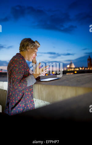 Young businesswoman using digital tablet tout en bavardant sur smartphone la nuit, Londres, UK Banque D'Images