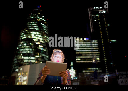 Femme mature en face d'immeubles de bureaux using digital tablet at night, London, UK Banque D'Images