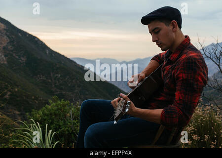 Jeune homme assis dans un paysage de montagne portant guitare acoustique, Los Angeles, Californie, USA Banque D'Images