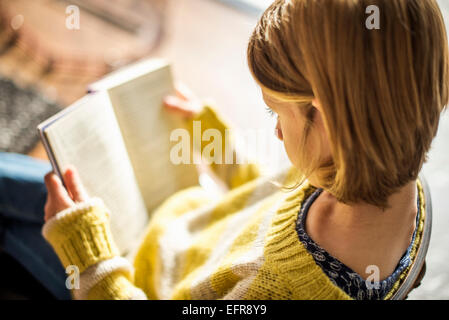 Portrait d'une fille blonde dans un cavalier jaune assis sur une chaise, lisant un livre. Banque D'Images