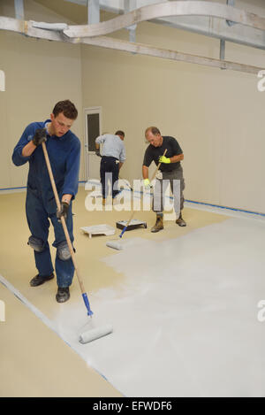 Trois commerçants roll une seconde couche d'époxy teintée produit sur le plancher d'un bâtiment industriel Banque D'Images