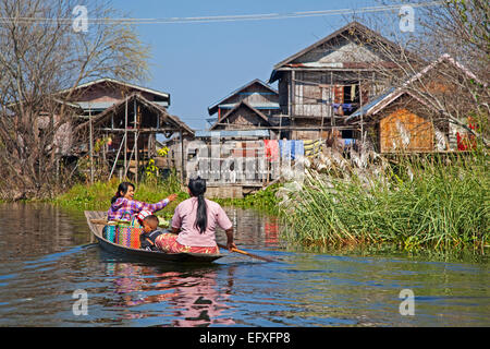 Les femmes avec enfant ethnie Intha en proa à aviron guindé village avec les maisons en bois sur pilotis au Lac Inle, Nyaungshwe, Myanmar / Birmanie Banque D'Images