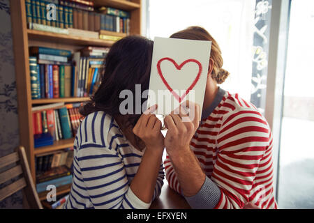 Flirty couple sitting in library derrière le papier avec coeur rouge Banque D'Images