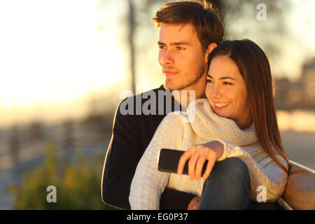 Couple et s'étreindre à au coucher du soleil assis sur un banc et la tenue d'un téléphone intelligent Banque D'Images