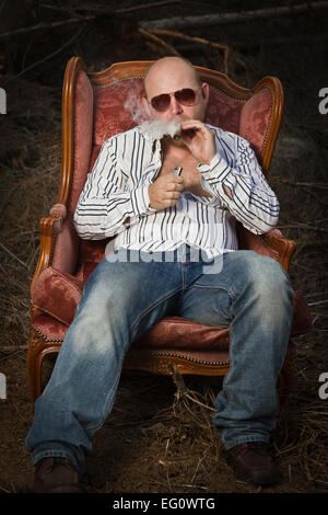 L'homme louche / dude dans un classique vintage chaise au milieu d'une zone d'exploitation forestière. Le feu vers le haut et la fumée d'un cigare et boire du Cognac dans w Banque D'Images