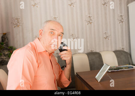 Close up surpris vieil homme de sa barbe à l'aide de rasoir électrique à sa table à l'intérieur de sa maison en regardant la caméra. Banque D'Images