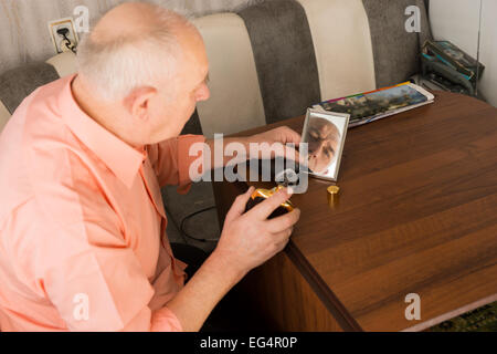 Close up assis vieux homme chauve avec l'après-rasage bouteille sur sa main à la recherche au petit miroir sur table en bois. Banque D'Images