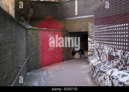 Femme sans-abri assis sous le couvert à un passage souterrain dans la ville de Londres, Royaume-Uni. Assis blottis et enveloppé du froid avec ses sacs d'effets personnels. Un panneau rouge peint sur le mur dans la forme d'une flèche indique surveillez cet espace. Banque D'Images