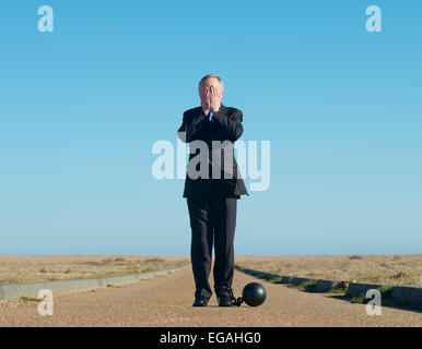 Homme d'affaires en costume sombre, attaché à une boule et chaîne avec sa tête dans ses mains. Il se dresse au milieu d'une route à distance. Banque D'Images