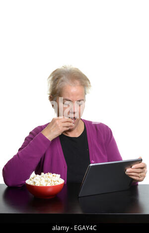 Femme d'âge moyen à l'aide d'un ordinateur tablette tandis que eating popcorn at home Banque D'Images