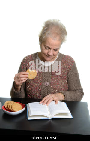 Femme d'âge moyen de la lecture d'un livre tout en mangeant des cookies à la maison Banque D'Images