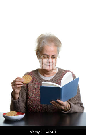 Femme d'âge moyen de la lecture d'un livre tout en mangeant des cookies à la maison Banque D'Images