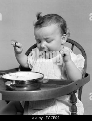 1940 BABY GIRL de manger des aliments dans une chaise haute STICKING OUT TONGUE Banque D'Images