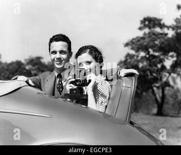 1930 COUPLE SITTING IN JUMP SEAT OF CAR pointant à l'APPAREIL PHOTO VIEWER Banque D'Images