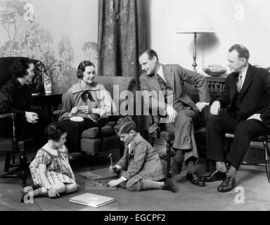 1930 DANS LA SALLE DE SÉJOUR FAMILLE Garçon Fille JOUANT SUR LE PLANCHER ALORS QUE Mère et père PARLER AVEC UN AUTRE COUPLE Banque D'Images