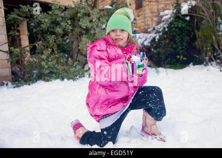 Petite fille mignonne debout sur un genou dans la neige avec une boule de neige dans ses mains Banque D'Images