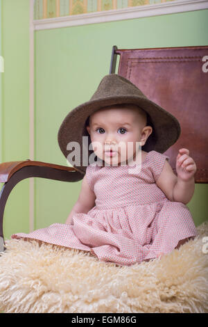 Mignon bébé portant une longue robe et chapeau de la mère. Assis sur une chaise dans la chambre Banque D'Images