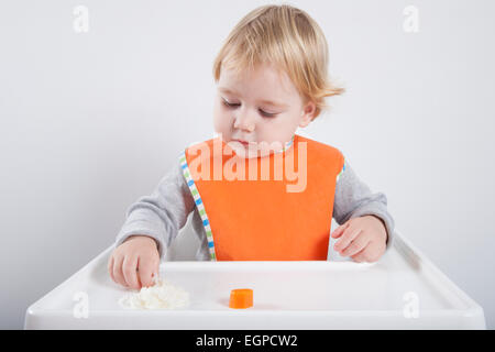 Blond caucasian baby 17 mois bavoir orange chandail gris repas manger avec sa main en blanc chaise haute Banque D'Images