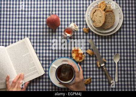 Femme ayant le petit déjeuner, ses mains et un livre, avec du pain et des bonbons sur nappe à carreaux Banque D'Images