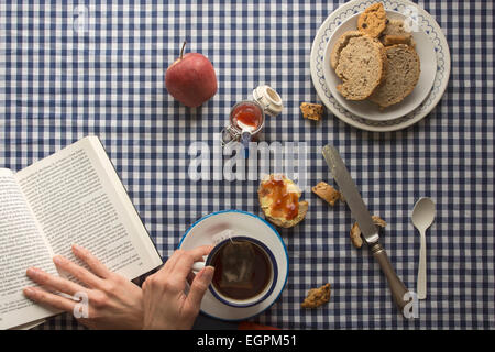 Femme ayant le petit déjeuner, ses mains et un livre, avec du pain et des bonbons sur nappe à carreaux Banque D'Images