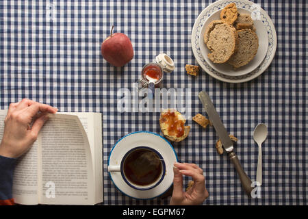 Femme ayant le petit déjeuner, ses mains et un livre, avec du pain et des bonbons sur nappe à carreaux Banque D'Images