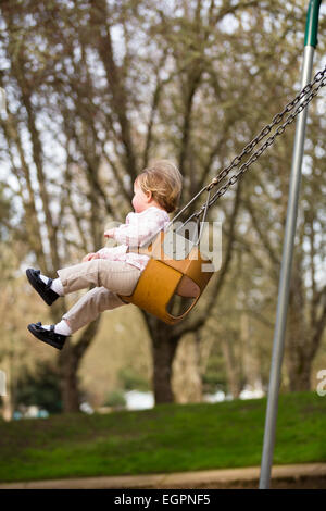 Un an fille sur une balançoire dans un parc dans la lumière naturelle. Banque D'Images