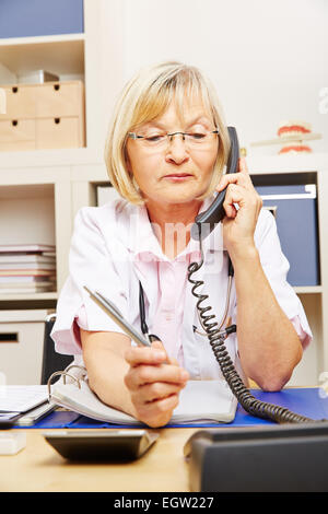 Vieille femme médecin sur appel assis dans son bureau au téléphone Banque D'Images