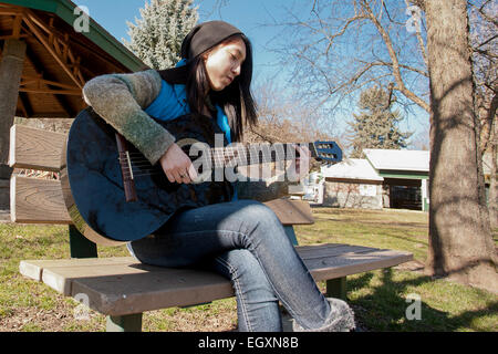 Un adolescent adultes se trouve sur un banc dans un parc et joue de la guitare. Banque D'Images