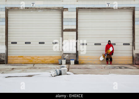 L'homme se tient debout devant une porte de garage en maintenant son snowboard. Banque D'Images