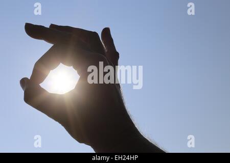 La main d'un homme rendant le symbole OK autour du soleil sur un fond de ciel bleu au-dessus de lui Banque D'Images