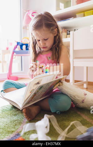 Petite fille assise sur le sol de sa chambre, lire un livre et en regardant son bracelet Banque D'Images