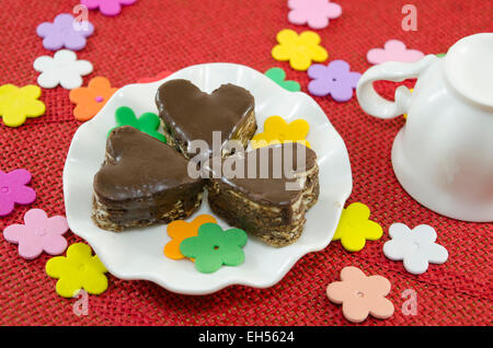 Les cookies au chocolat en forme de coeur sur une assiette décorée de fleurs en papier sur une nappe rouge Banque D'Images