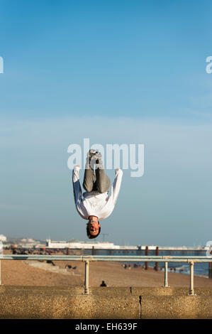 Un jeune homme fait des culbutes et retourne en arrière dans l'air alors qu'un groupe de jeunes font l'essentiel de la première journée de beau temps de printemps pratiquer l'art du parkour, free-sauter et faire des sauts périlleux, au large de la jetée sur le front de mer de Brighton à Brighton Pier en arrière-plan. Brighton, East Sussex, Angleterre, Royaume-Uni. Banque D'Images